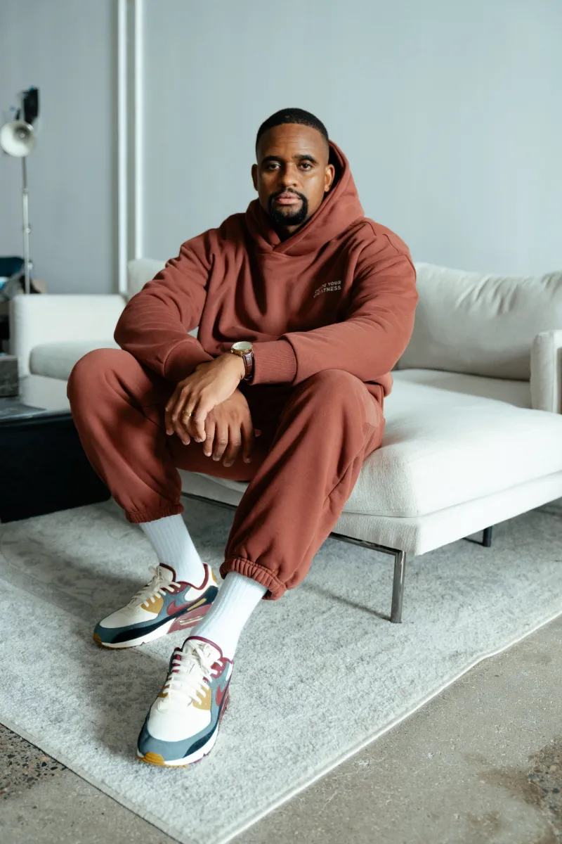 Man seated on a white couch wearing the Signature Sweatpants and Signature Boxy Hoodie in brown