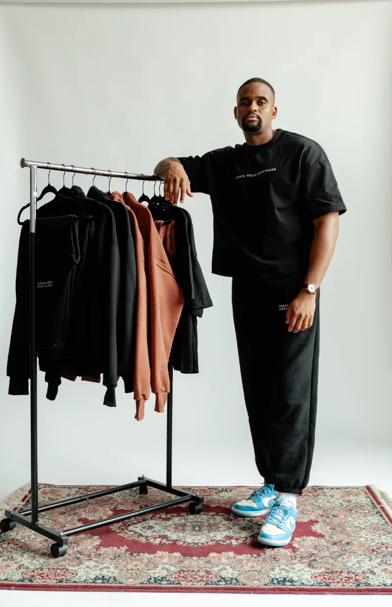 Man in a black Create Your Greatness tee and sweatpants leaning on a clothing rail of black and brown pieces